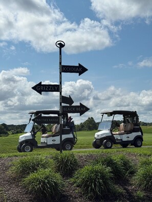 Two golf carts parked on a grassy field with directional signs pointing to Grizzle, Kodiak, and Black Bear. - Pond View Bear Trap Home- 4BR 3.5BA - Screened Porch - Near Bethany Beach & Golf (Ocean View)