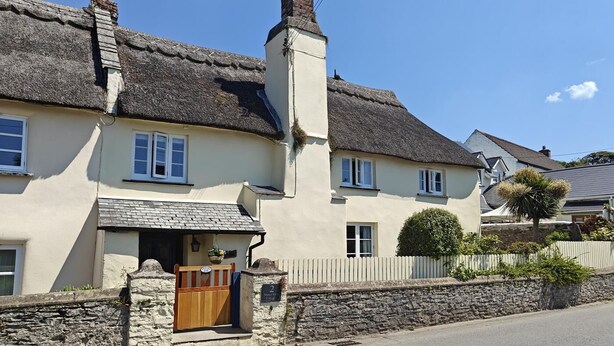 Une maison de campagne traditionnelle, avec un toit de chaume, un mur en pierre, un portail en bois et une enseigne indiquant qu’il s’agit d’une maison d’hôtes.