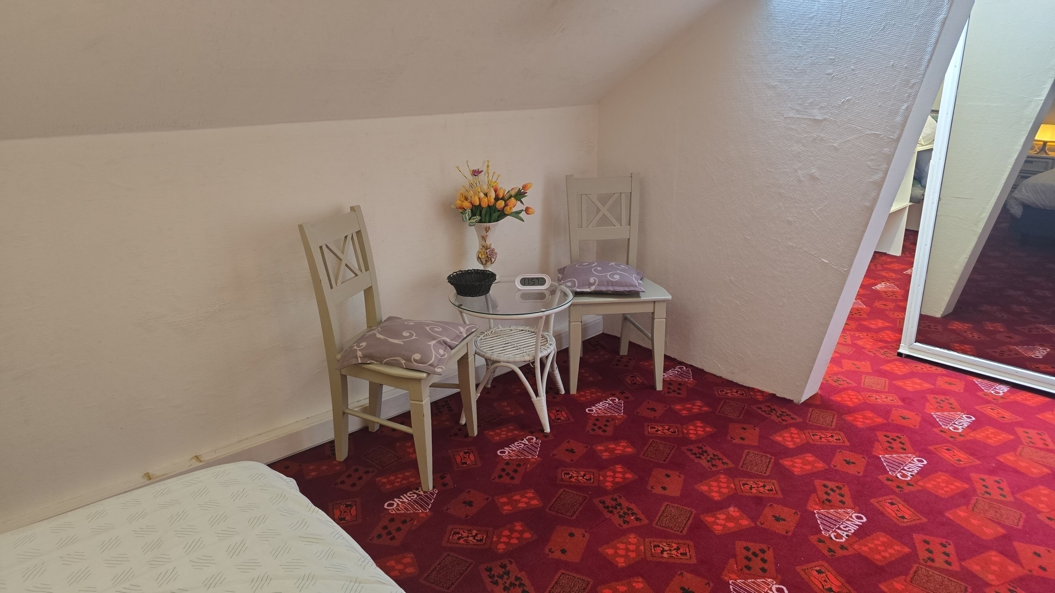 Une petite pièce comprenant un lit, une table avec des chaises, un vase avec des fleurs et un tapis rouge à motifs.