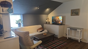 Ein Wohnzimmer mit einer Couch, einem Couchtisch, einem Fernseher auf einem Schrank, einem kleinen Tisch, einer Zimmerpflanze und einem Bild an der Wand.