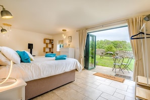 A bedroom with a bed, bedside lamps, a dresser, a mirror, and a view of a garden through a glass door. - 3 Bed in Duntisbourne Abbots (oc-f42787) (Duntisbourne Abbots)