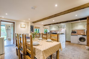 A dining area with a table set for four, wooden chairs, a vase with flowers, and a view of the kitchen with a washing machine. - 3 Bed in Duntisbourne Abbots (oc-f42787) (Duntisbourne Abbots)