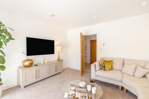 Ein modernes Wohnzimmer mit einem Flachbildfernseher, einem beige farbenen Sofa, einem Glastisch und einem schlichten Schrank.