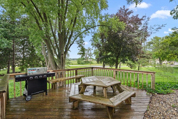 Una terraza de madera con una parrilla y una mesa de picnic, rodeada de árboles y vistas a un campo de golf.