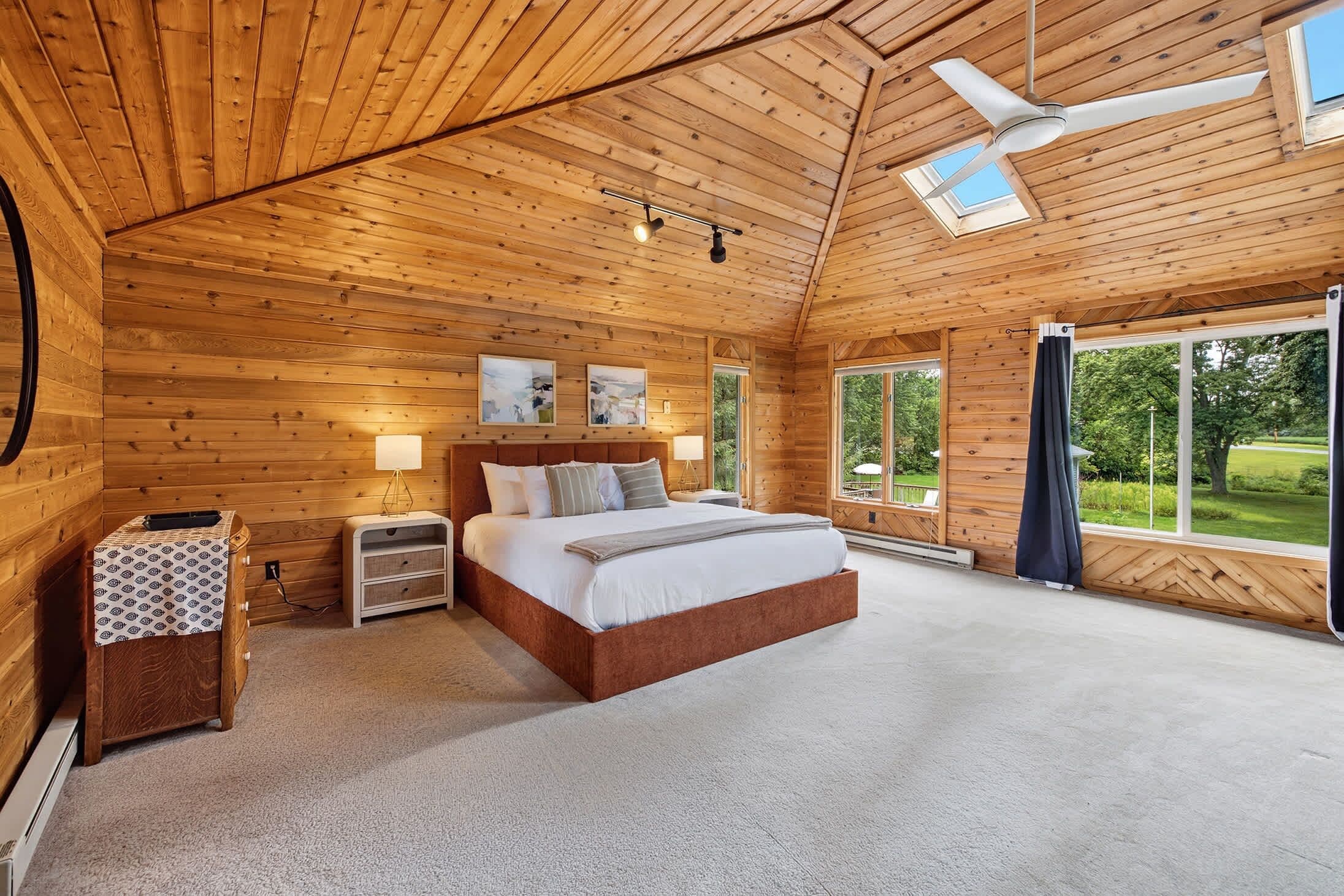 A bedroom with a wooden ceiling, a bed, a nightstand, a lamp, and a view of the outdoors.