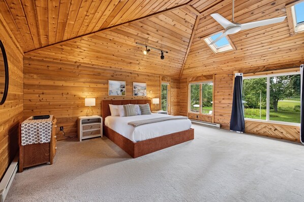 A bedroom with a wooden ceiling, a bed, a nightstand, a lamp, and a view of the outdoors.