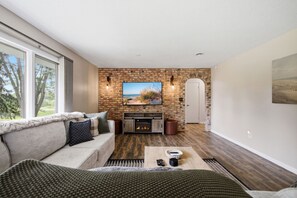 A living room with a fireplace, a television mounted on a brick wall, and a large window overlooking a natural landscape.