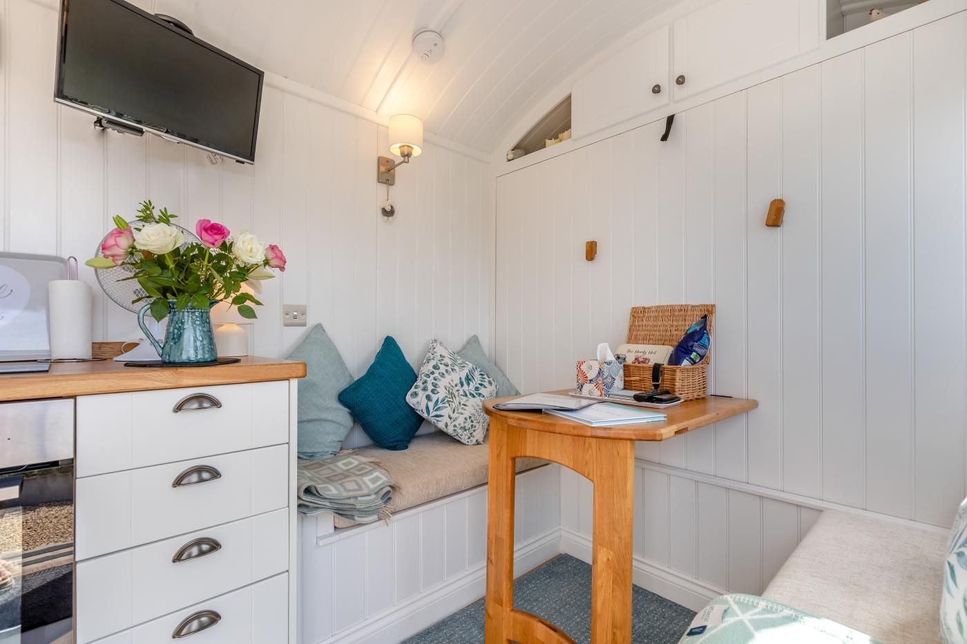 A cozy room with a wooden desk, a comfortable seating area with cushions, a television mounted on the wall, and a small table with a basket.