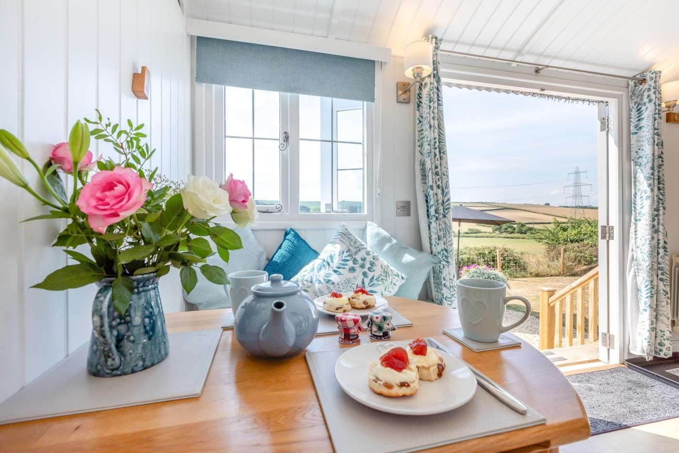 A room with a wooden table, a vase with flowers, a kettle, and a plate with food.