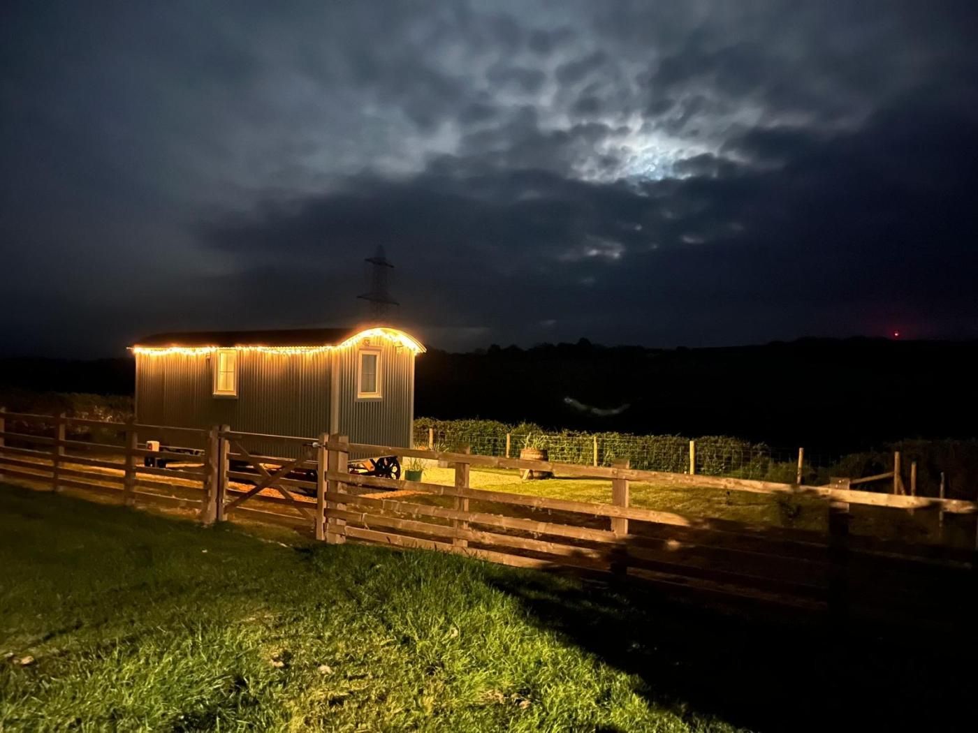A small, illuminated structure with a sloped roof, a door, and a window, surrounded by a wooden fence and grass.