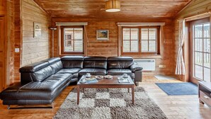 Ein Holzblockhaus-Interieur mit einem ledernen Ecksofa, einem Holztisch und einem großen Fenster mit Fensterläden.