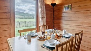 Eine Holzhütte mit einem für das Frühstück gedeckten Esstisch und Blick auf eine grüne Landschaft, vor der eine Holzlattenzaun steht.