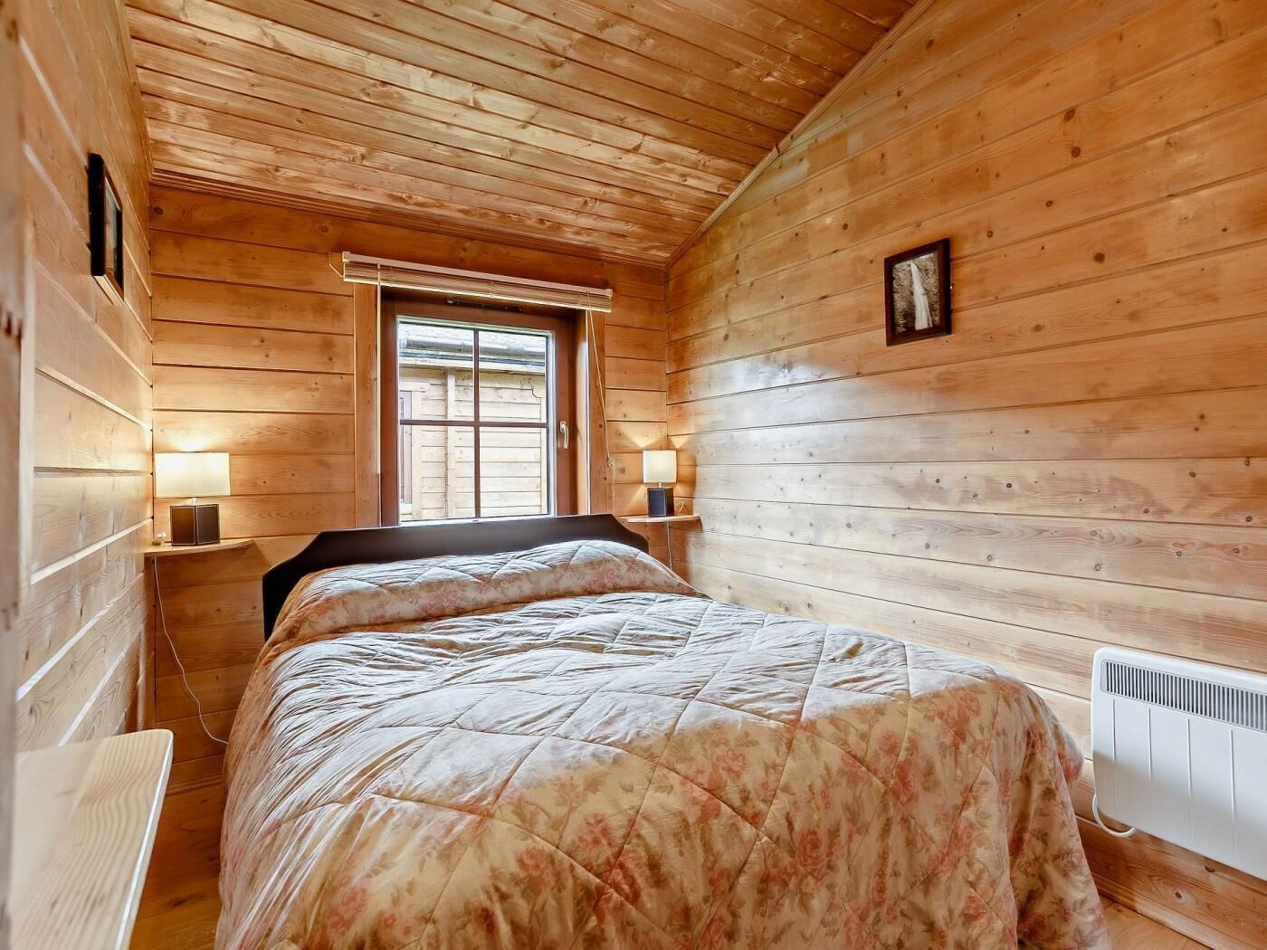 Ett sovrum i en stuga med träpanel, inrett med en säng, två sänglampor, ett fönster och ett element.