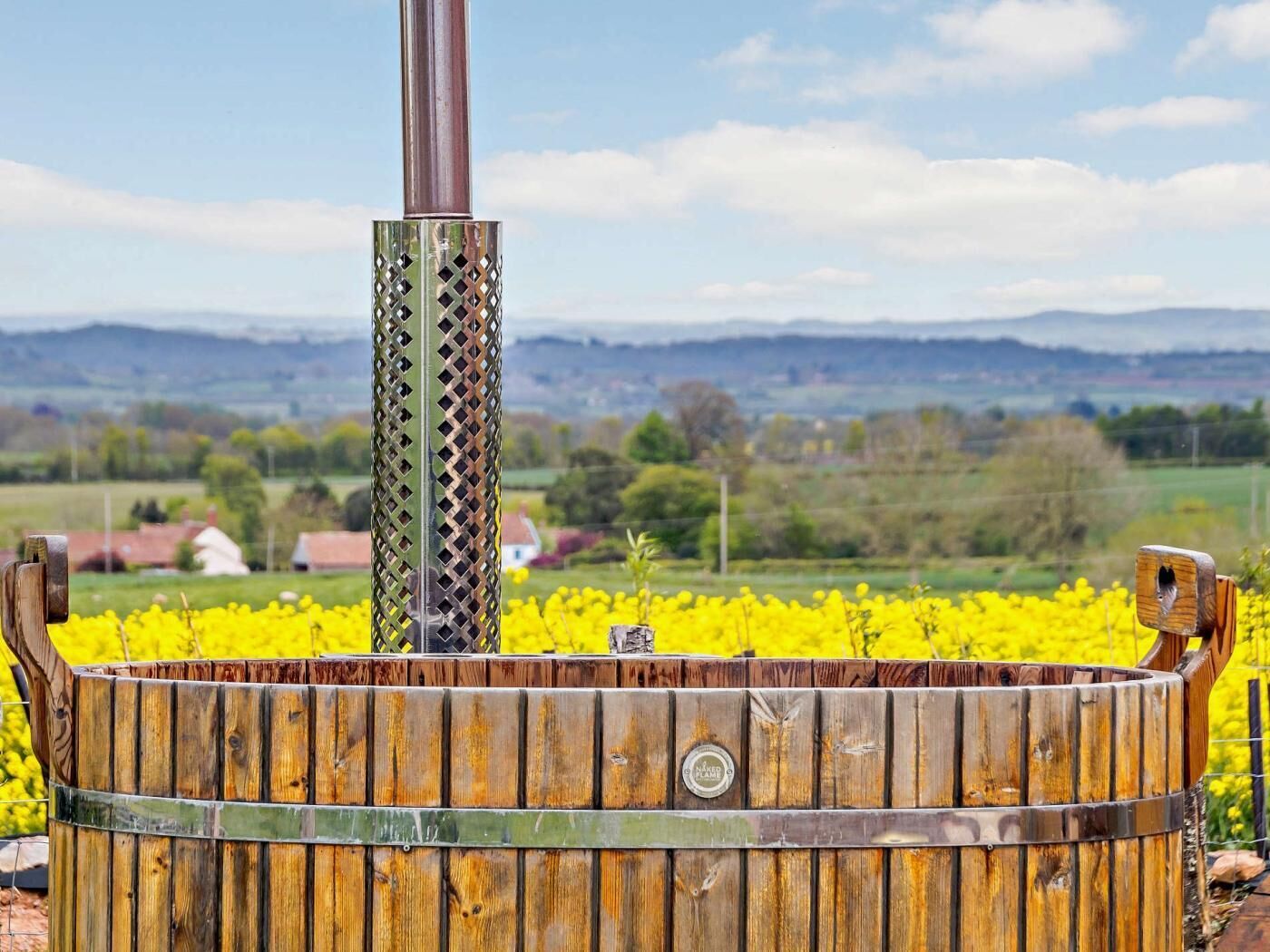 A wooden barrel with a metal chimney, set against a rural landscape with fields and distant hills.