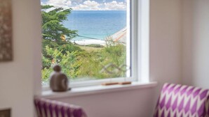 Ein Fenster mit Blick auf Strand und Meer, eine kleine Statue im Fensterbrett und ein violett gemustertes Stuhl.