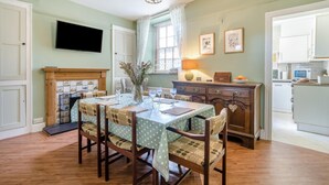 Un coin repas avec une table dressée pour quatre personnes, une cheminée et une cuisine visible à travers une porte ouverte.