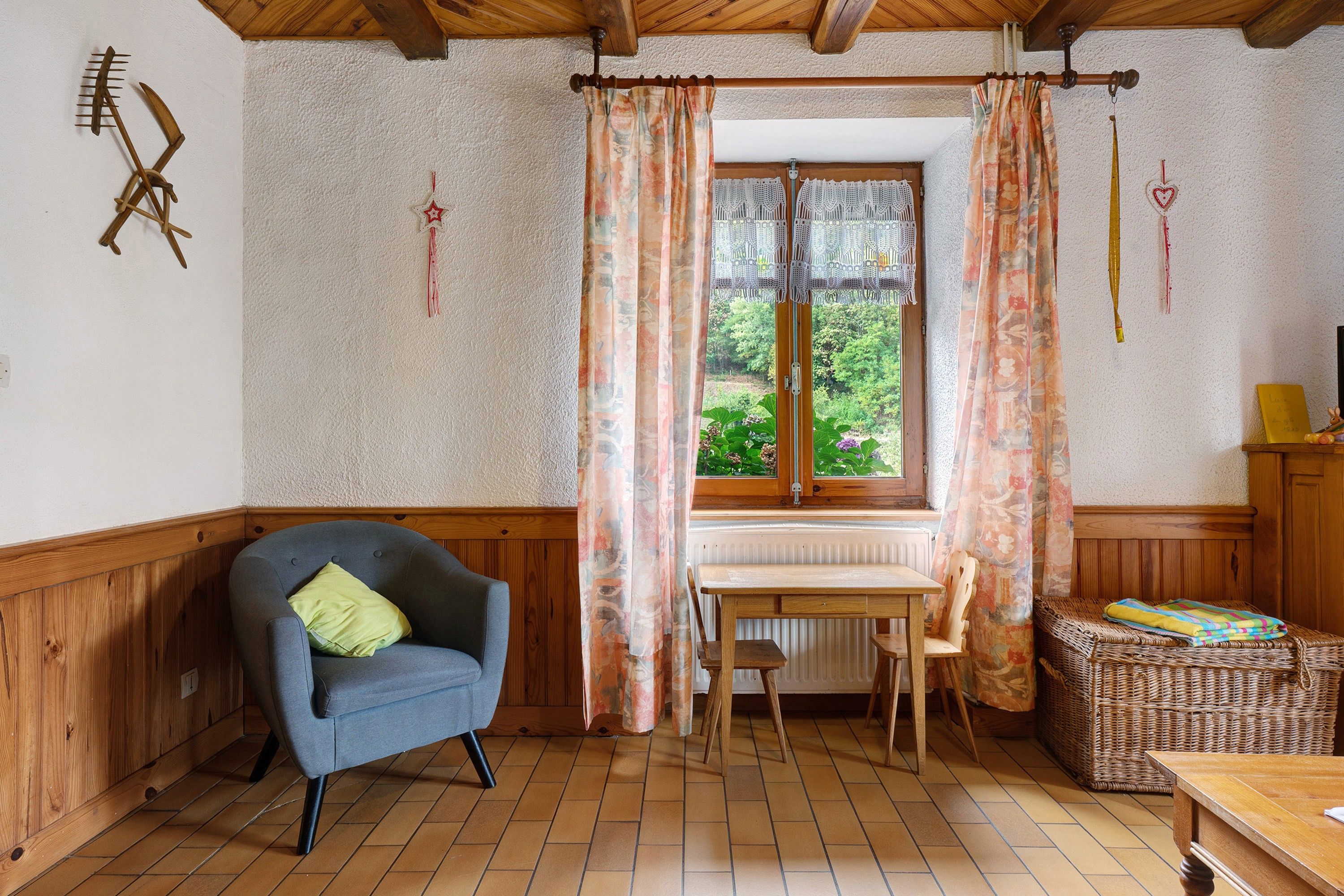 Une pièce chaleureuse avec un plafond en bois, un fauteuil bleu, une table en bois et une fenêtre ornée de rideaux à motifs floraux.