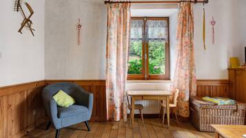 Une pièce chaleureuse avec un plafond en bois, un fauteuil bleu, une table en bois et une fenêtre ornée de rideaux à motifs floraux.