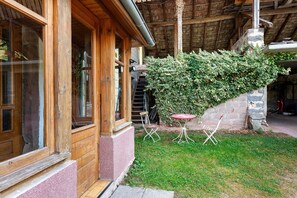 A wooden cabin with a glass door, a small outdoor seating area, and a stone wall covered with ivy.