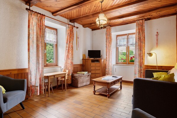 A cozy living room with a wooden ceiling, a television, a table with a remote, and a window with curtains.