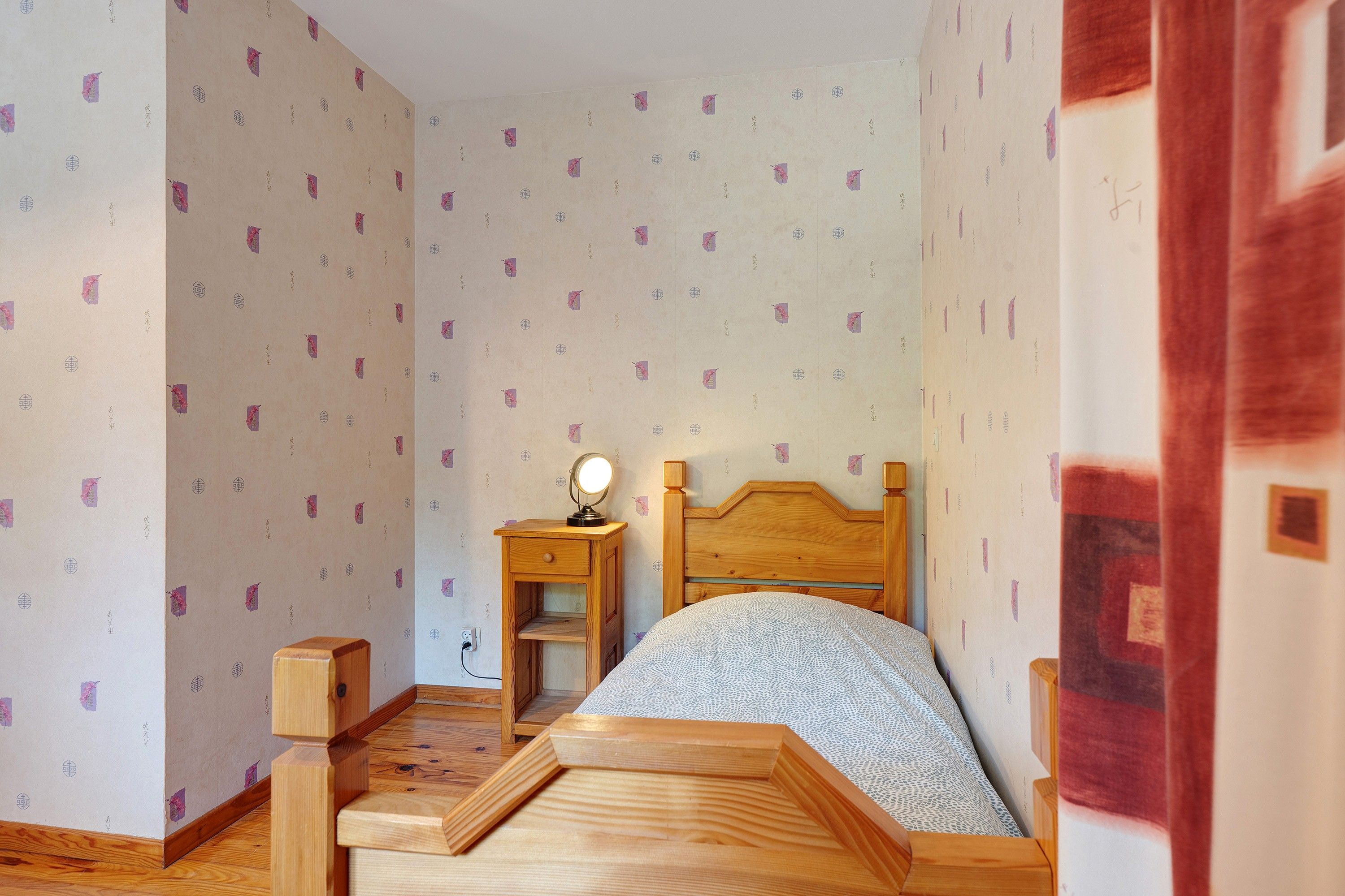Een slaapkamer met een houten bed, een nachtkastje met een lamp en behang met een patroon.