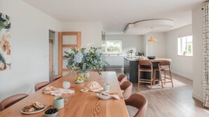 A modern kitchen with a central island, a dining area with a long wooden table, and a view of the adjacent living space. - 5 Bed in St. Minver (oc-l50188) (Saint Minver)