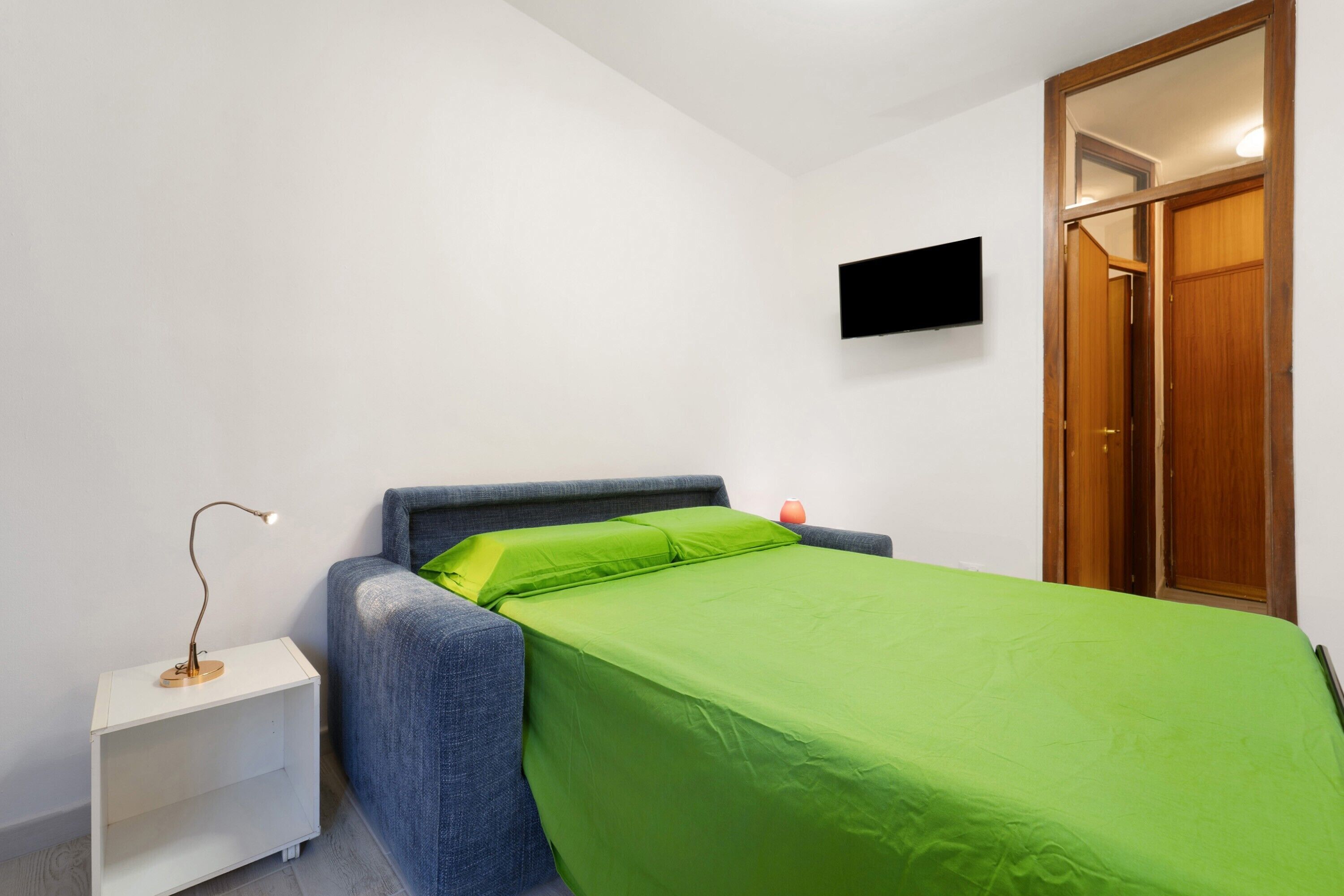Een slaapkamer met een bed, nachtkastje, lamp, televisie aan de muur en een houten garderobe.