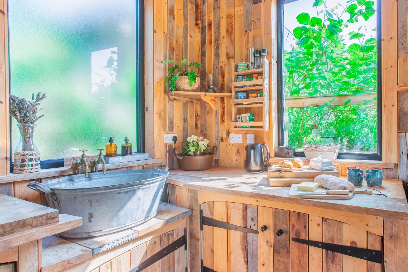 Een landelijke keuken met houten kastjes, een grote metalen spoelbak en een raam met uitzicht op groen.
