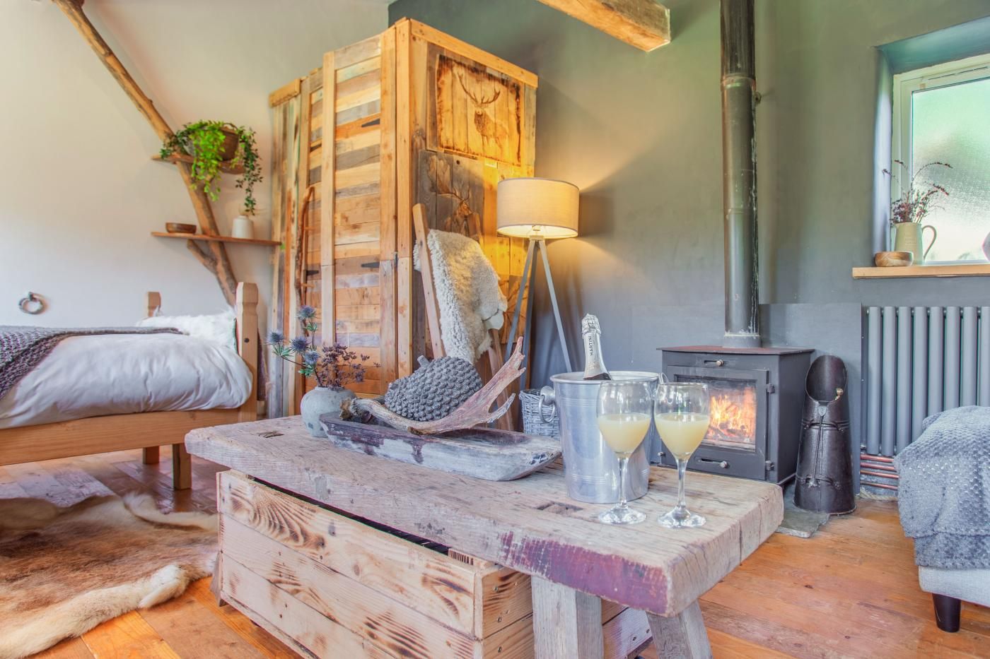 A cozy room with a bed, a wooden cabinet, a lamp, a fireplace, and a rustic dining table with glasses.
