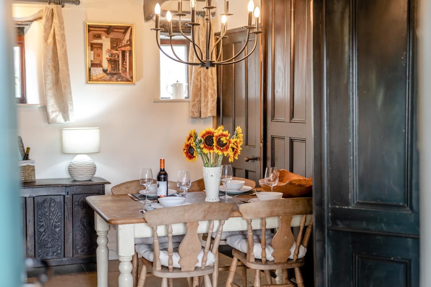 A dining area with a wooden table, chairs, a chandelier, and a vase with sunflowers.
