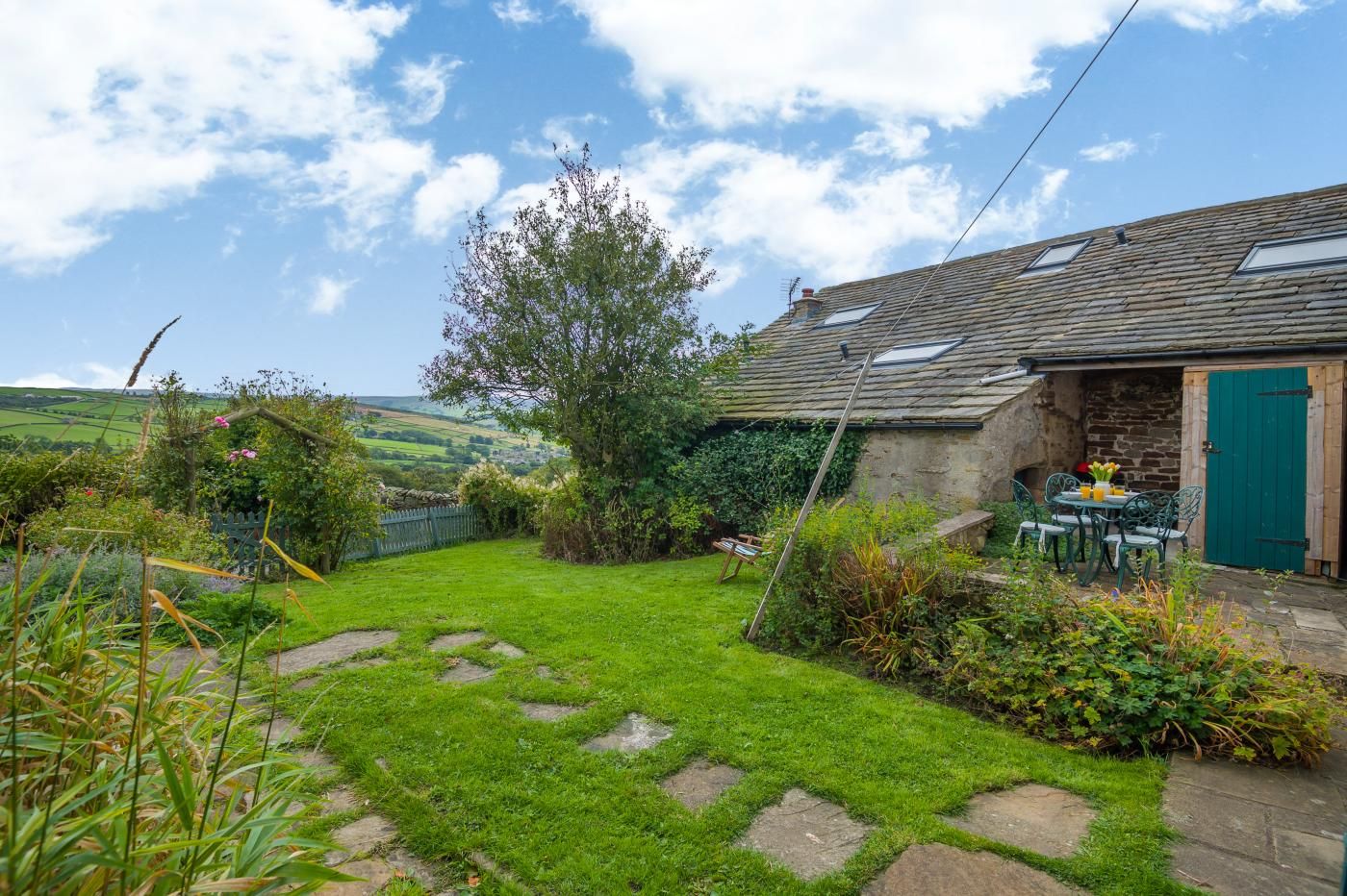 A rustic stone house with a green door, a garden with a stone path, and a view of the countryside.