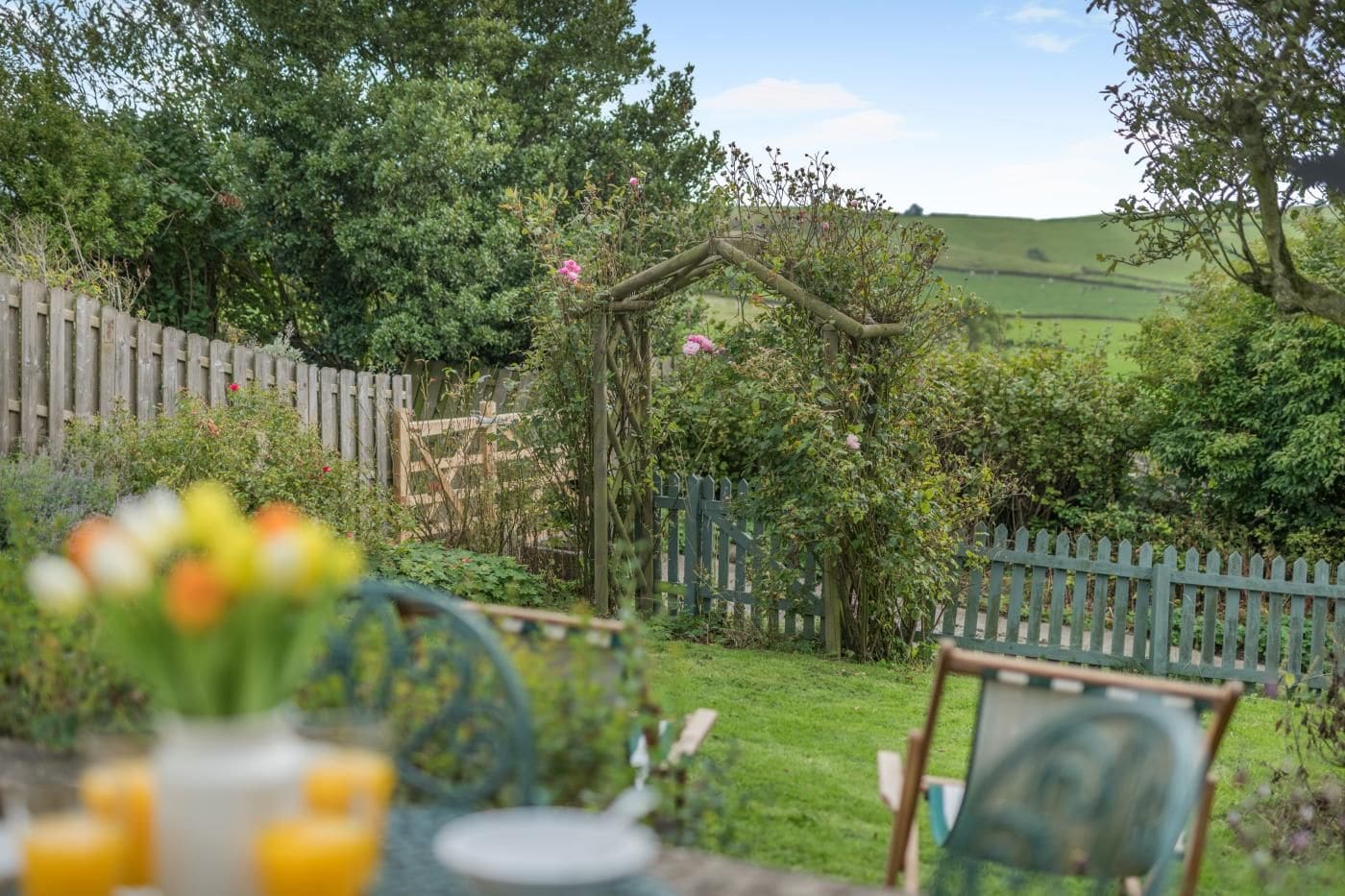 A garden with a wooden fence, a pergola with climbing plants, and a table with chairs in the foreground.
