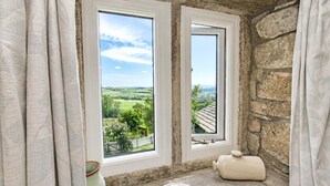Quarto com paredes de pedra e duas janelas com vista para uma paisagem rural, uma pequena mesa com um bule e um prato decorativo.