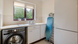 Ett kompakt kök med tvättmaskin, diskho, ett fönster med utsikt över grönområden och en blå strykjärnsbräda.
