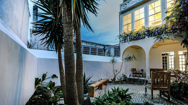 A courtyard with a palm tree, benches, and a building with arched windows and a balcony with purple flowers. - GuestReady - Doctor House Gardens (Horta)