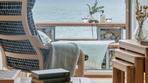 Ett rum med utsikt över havet, en stol, ett bord med böcker och en liten växt på fönsterbrädan.