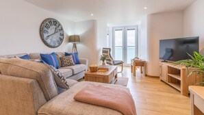 A living room with a sofa, a coffee table, a TV, a large clock, and a view through a glass door. - 2 Bed in Port Isaac (oc-l50168) (Port Isaac)