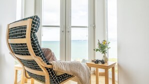 A room with a large window, a wooden chair with a patterned cushion, a small table with a vase and headphones, and a view of the sea. - 2 Bed in Port Isaac (oc-l50105) (Port Isaac)