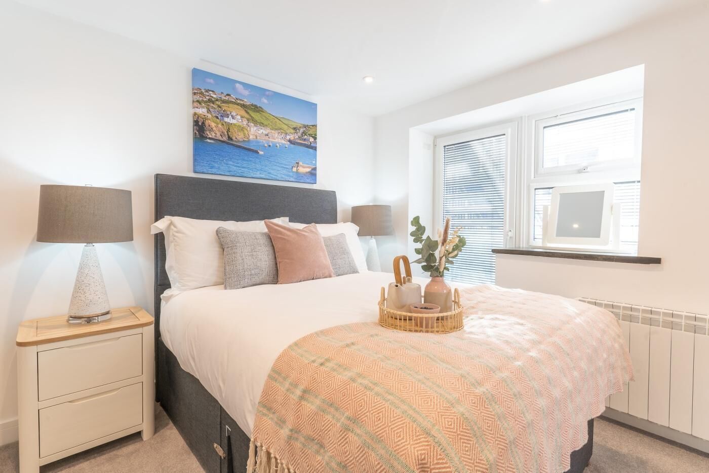 A bedroom with a bed, bedside lamps, a nightstand, a window with blinds, a wall-mounted picture, and a bedside table with a lamp and a vase with flowers.
