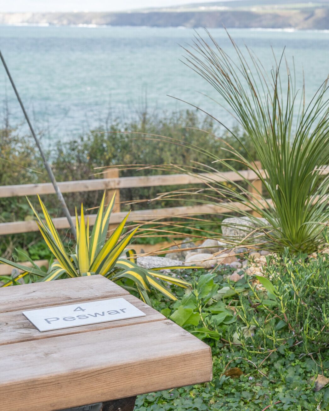 En træbænk med et skilt, der viser '4 Peswar', i en have med planter og udsigt til havet.