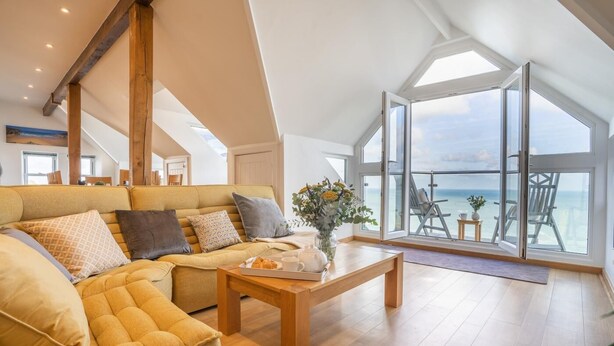 Uma sala de estar ampla, com teto alto, um sofá grande, uma mesa de centro e vista para o mar por grandes janelas.