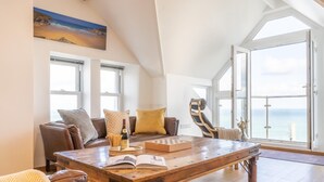 Uma sala ampla com uma mesa de madeira, um sofá e uma cadeira, uma janela grande com vista para o mar, e um quadro na parede.