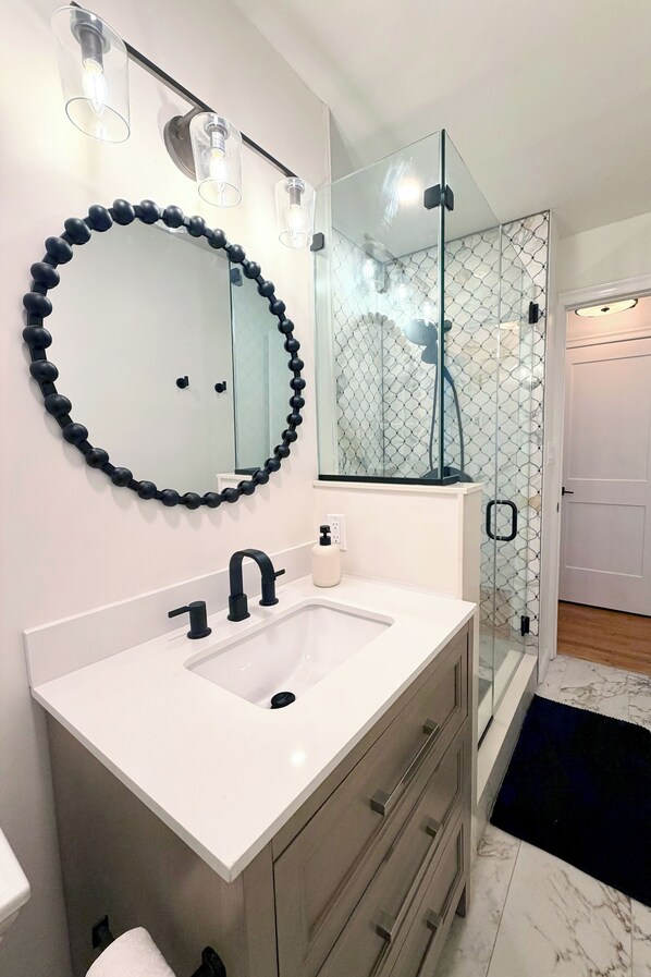 Un baño con lavamanos blanco, grifo negro y un espejo redondo adornado con un collar de cuentas negras.