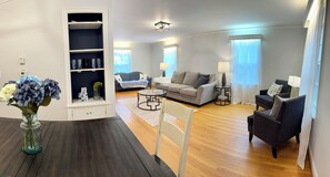A living room with a dark wooden floor, a glass coffee table, a grey sofa, a black armchair, and a white chair. There is a bookshelf with decorative items and a vase with flowers.