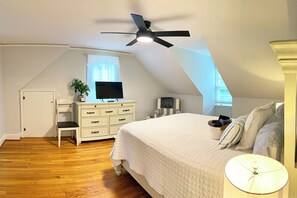 Een slaapkamer met een schuine plafond, een bed met een witte sprei, een televisie op een dressoir, een plant en een plafondventilator.