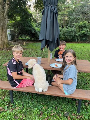 Un grupo de personas sentadas en una mesa de madera al aire libre, con un perro, rodeadas de follaje.