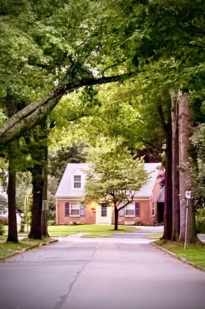 Una calle residencial con una casa de ladrillo, árboles grandes y un camino de entrada despejado.