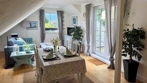 Ein gemütliches Zimmer mit Esstisch für zwei Personen, einer Couch, einem Fernseher und einem großen Fenster mit Vorhängen.