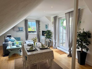 Ein gemütliches Zimmer mit Esstisch für zwei Personen, einer Couch, einem Fernseher und einem großen Fenster mit Vorhängen.
