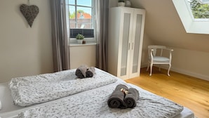 Ein Schlafzimmer mit zwei Betten, einem Fenster mit Vorhängen, einem weißen Kleiderschrank, einem weißen Sessel und einer herzförmigen Dekoration an der Wand.